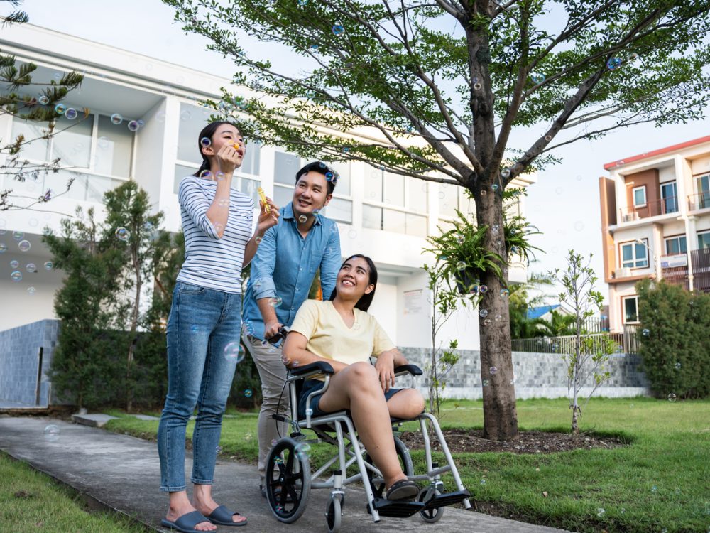 Group of Asian young man and woman friends walking together in garden. Attractive female legless amputee going outdoors on picnic with her friends with happiness during weekend activity in backyard.